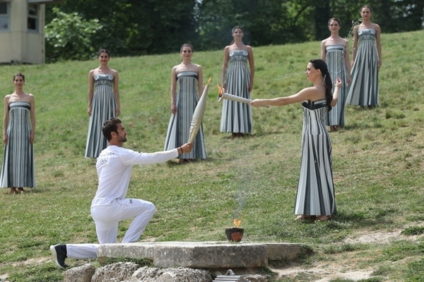 扮演最高女祭司的演員瑪麗·米娜（前右）點(diǎn)燃第一棒火炬手——希臘運(yùn)動(dòng)員、東京奧運(yùn)會(huì)男子賽艇項(xiàng)目金牌得主斯特凡諾斯·恩圖斯科斯手中的火炬。新華社記者 趙丁喆 攝